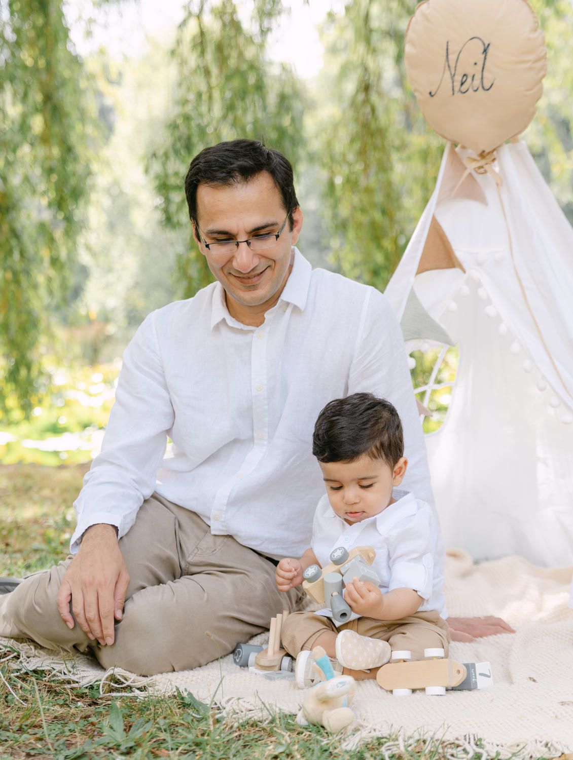 Father and toddler son sitting together on blanket outdoors near teepee tent with balloon at garden photoshoot.