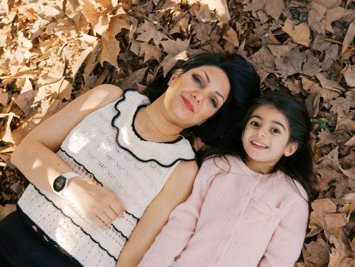 A mother and young daughter lying on autumn leaves, smiling up at the camera in a warm fall outdoor setting.