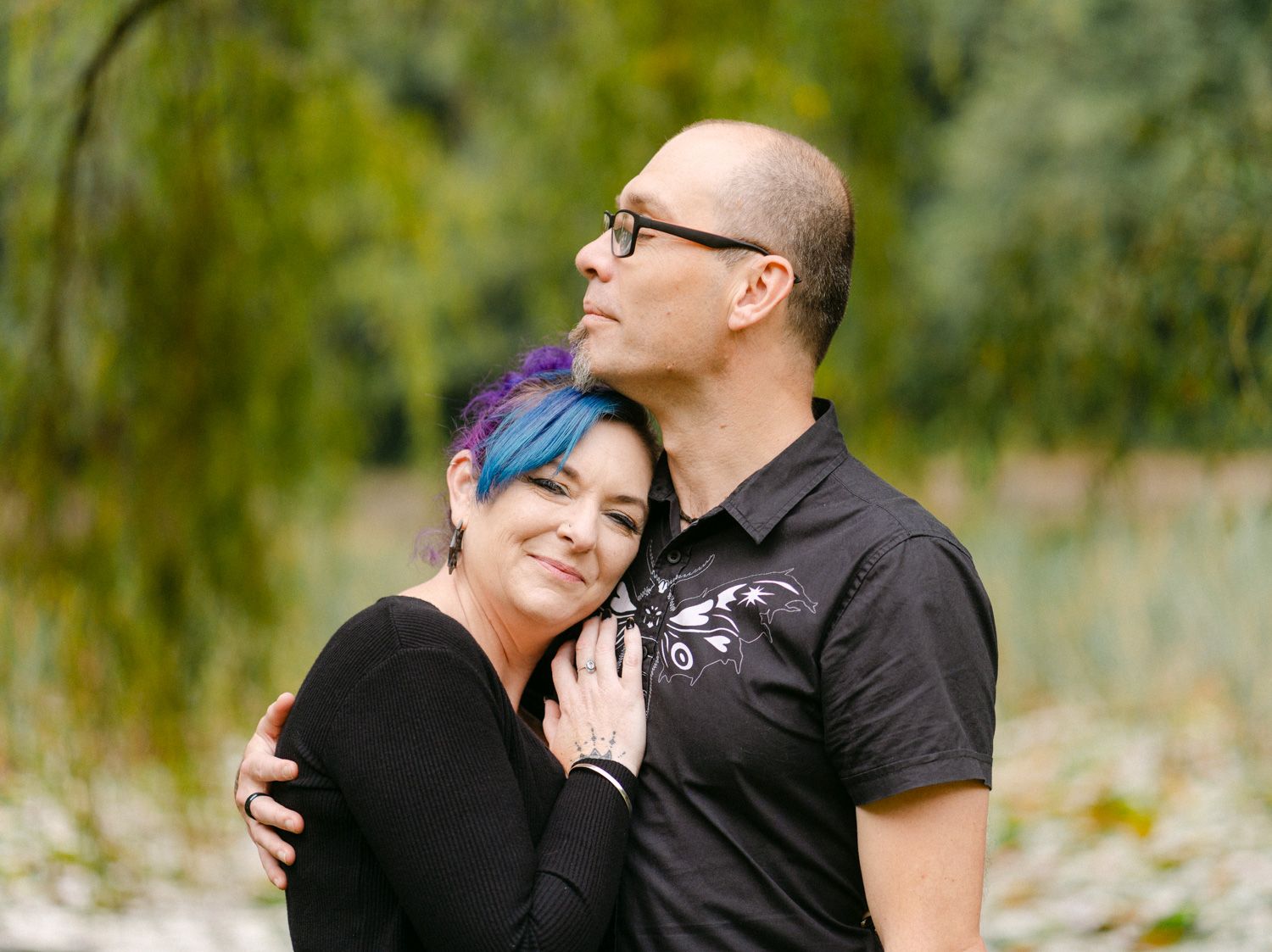 Couple embracing outdoors, woman with blue hair leaning on mans chest, surrounded by lush green trees.