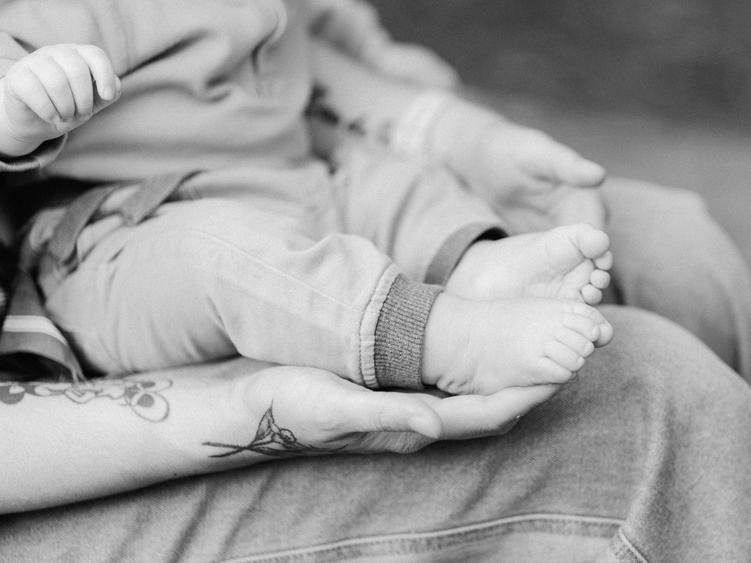 Black and white photo of a parent cradling a newborn baby's tiny feet in tattooed hands.