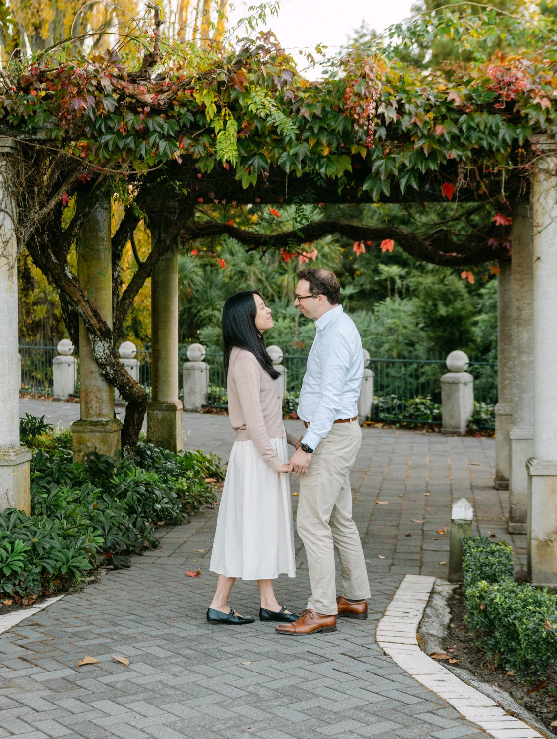 Couple holding hands facing each other under a vine-covered pergola in a lush garden during fall.