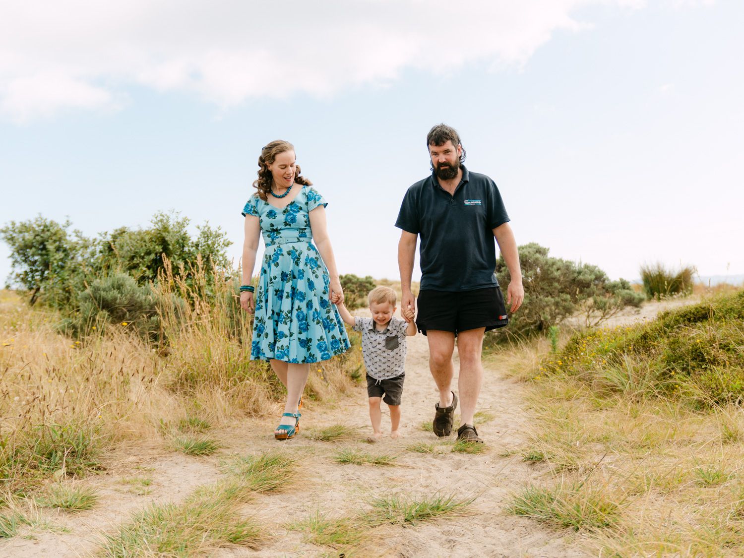 Family of three walking on a sandy nature trail, parents holding toddler's hands on a sunny day.