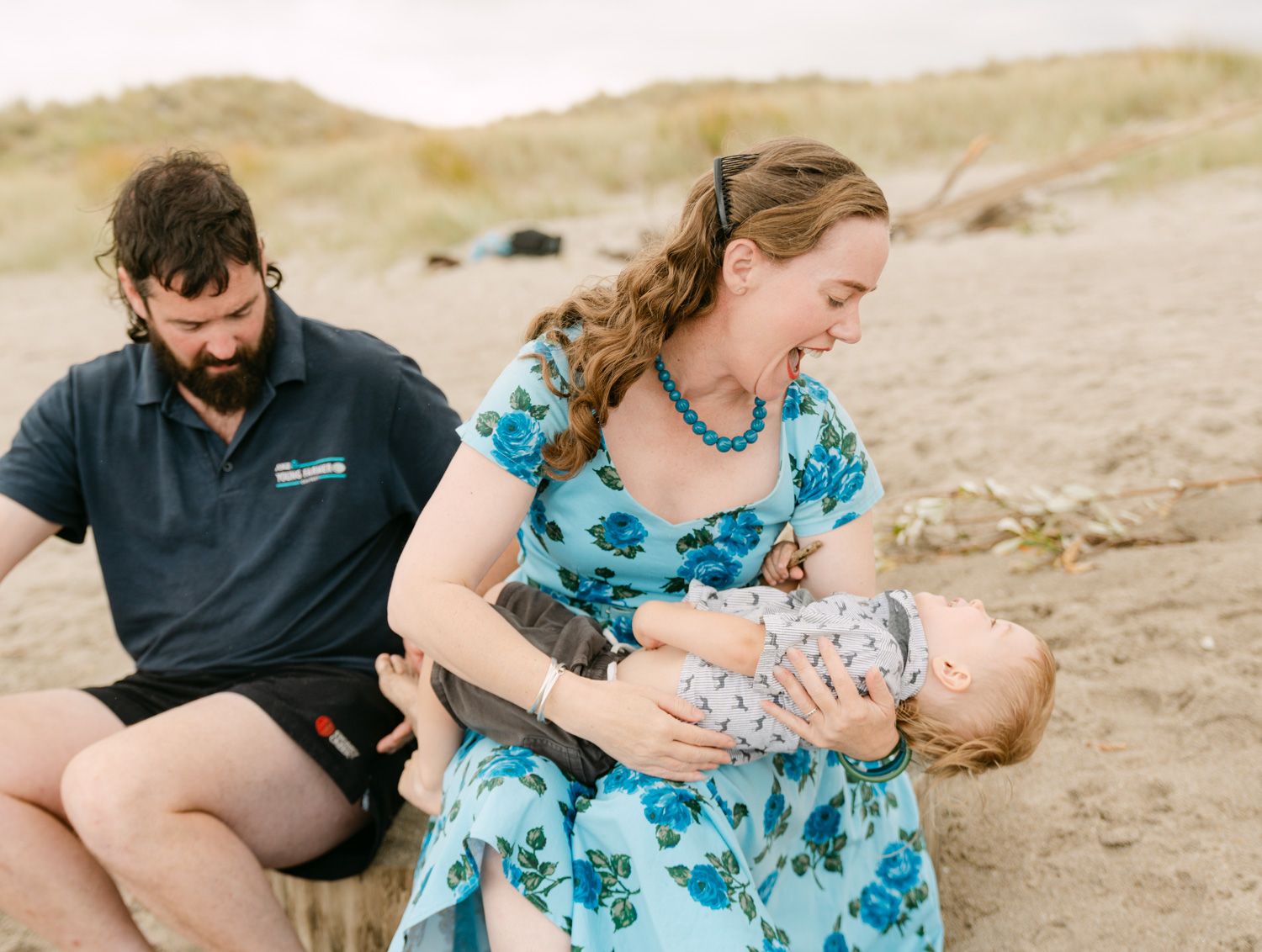Mother breastfeeding baby on sandy beach while father sits nearby, sharing a joyful family moment outdoors.