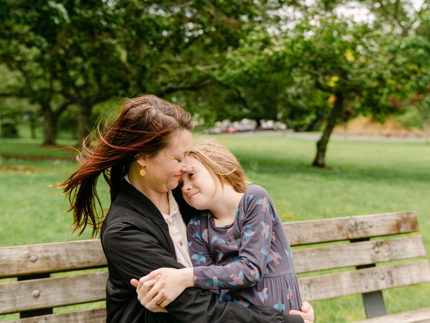 A mother and daughter share a joyful embrace on a park bench surrounded by lush green trees.