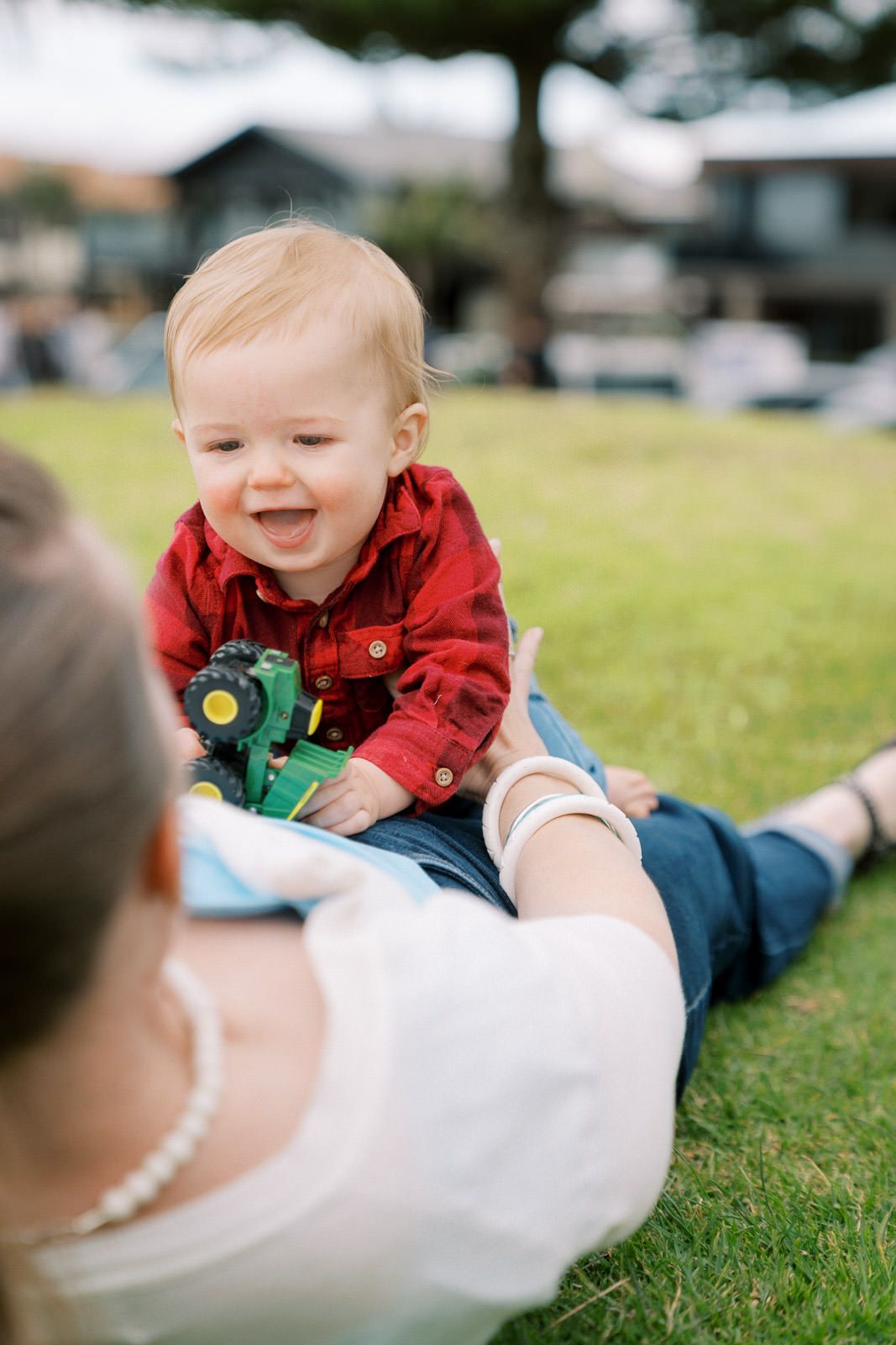 Happy baby in red plaid shirt laughing while holding green toy tractor, sitting with mother on grass outdoors.