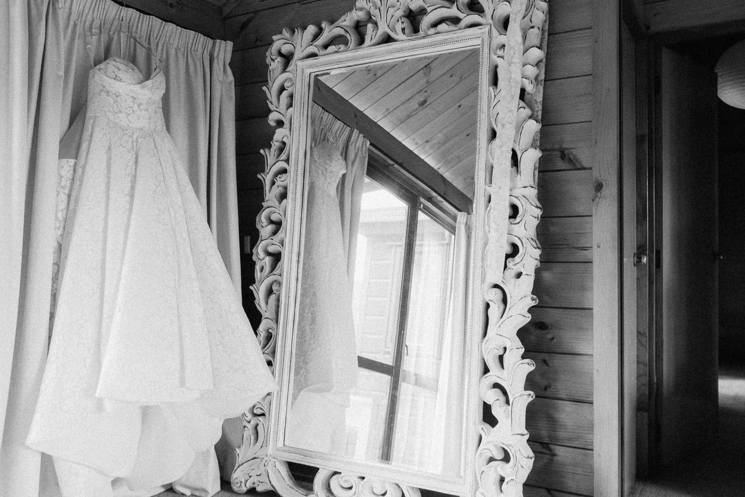 Black and white photo of a lace wedding dress hanging beside an ornate white mirror in a rustic wooden room.