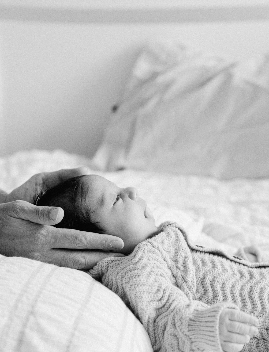 Black and white image of a parent's hand gently cradling a sleeping newborn on a white bed.