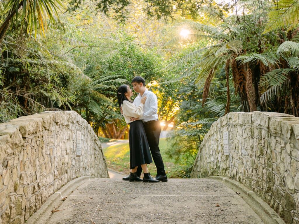 Couple sharing a romantic moment on a stone bridge surrounded by lush tropical ferns and trees at golden hour.