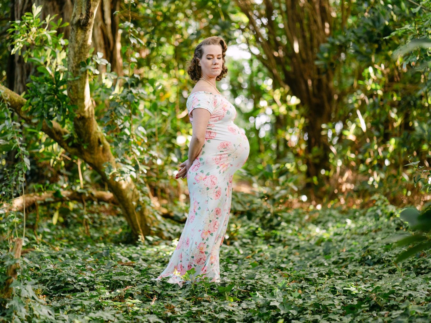 Pregnant woman in floral maxi dress standing in lush green forest, cradling baby bump during maternity photoshoot.