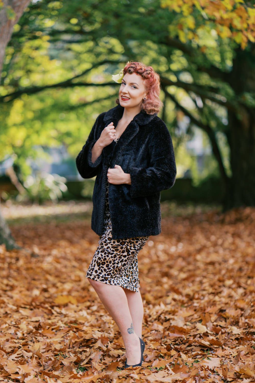 Woman in black faux fur coat and leopard print skirt posing among autumn leaves in a park.