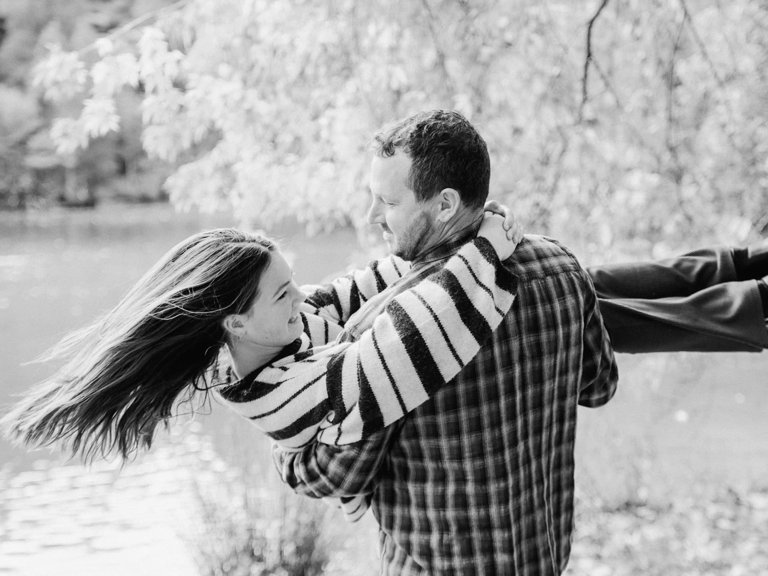 Black and white photo of a couple dancing and spinning outdoors near a lake with blossoming trees.