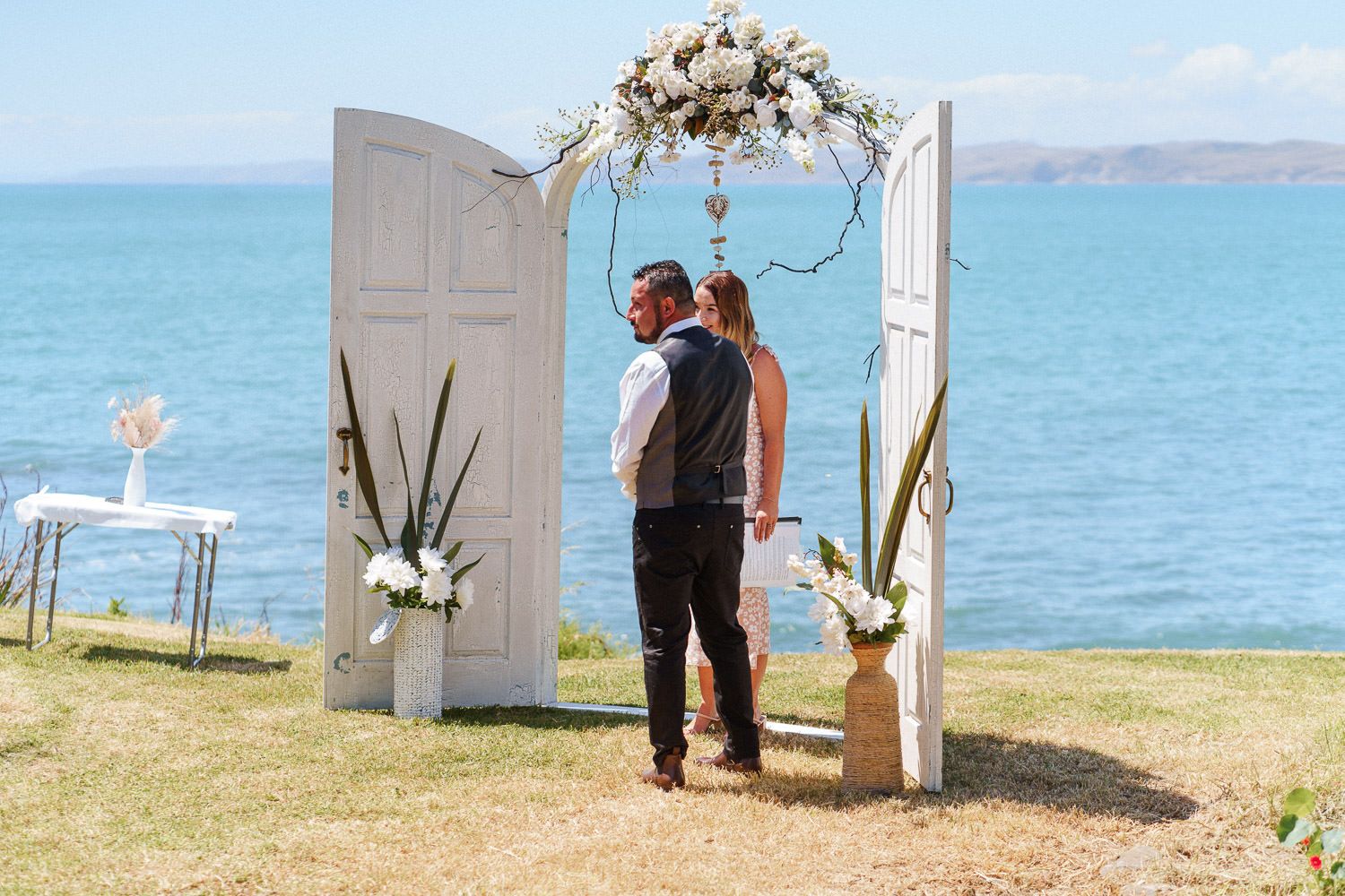 Couple kissing at outdoor wedding ceremony with white doors arch, floral decorations, and ocean backdrop.