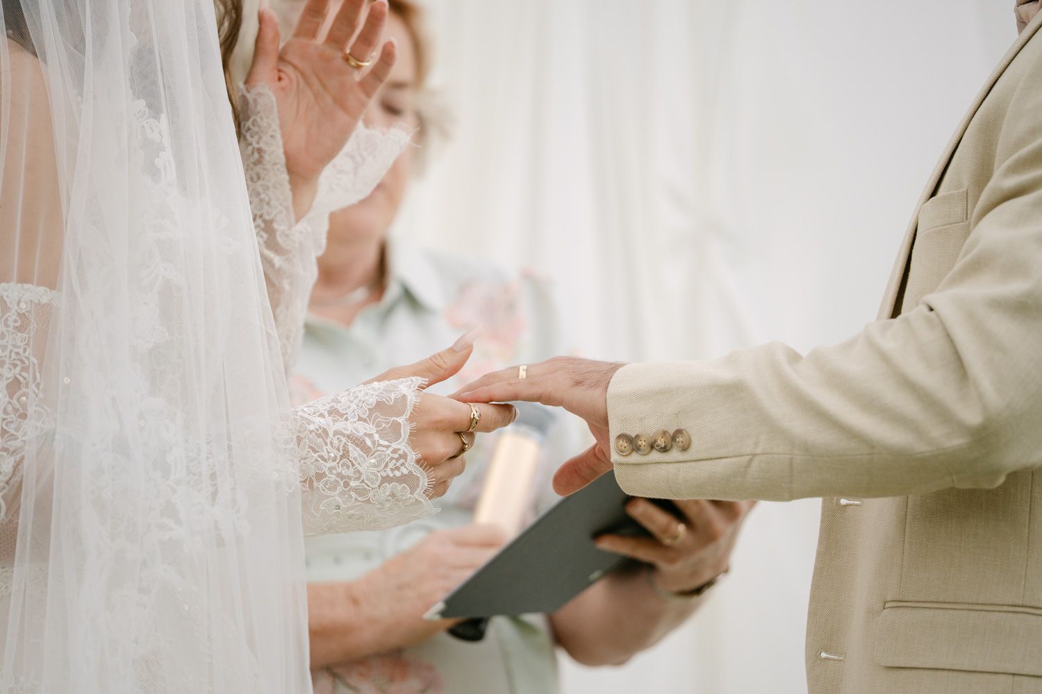 Bride and groom exchanging rings during wedding ceremony, officiant in background, lace bridal gown detail.