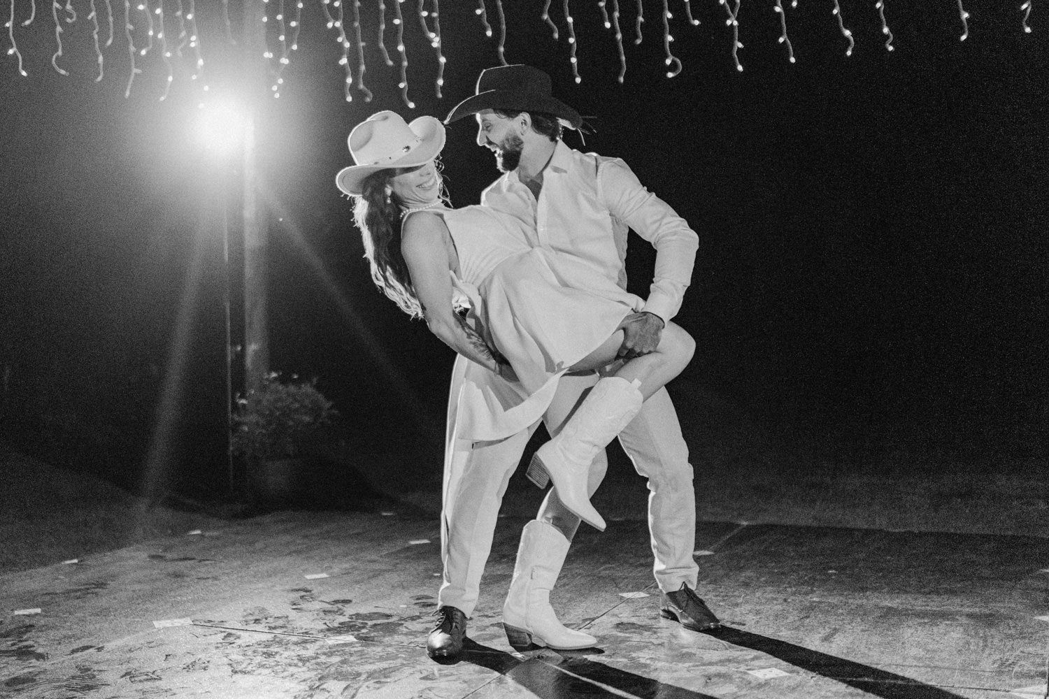 Black and white photo of a couple sharing a romantic dip kiss outdoors at night under twinkling string lights.