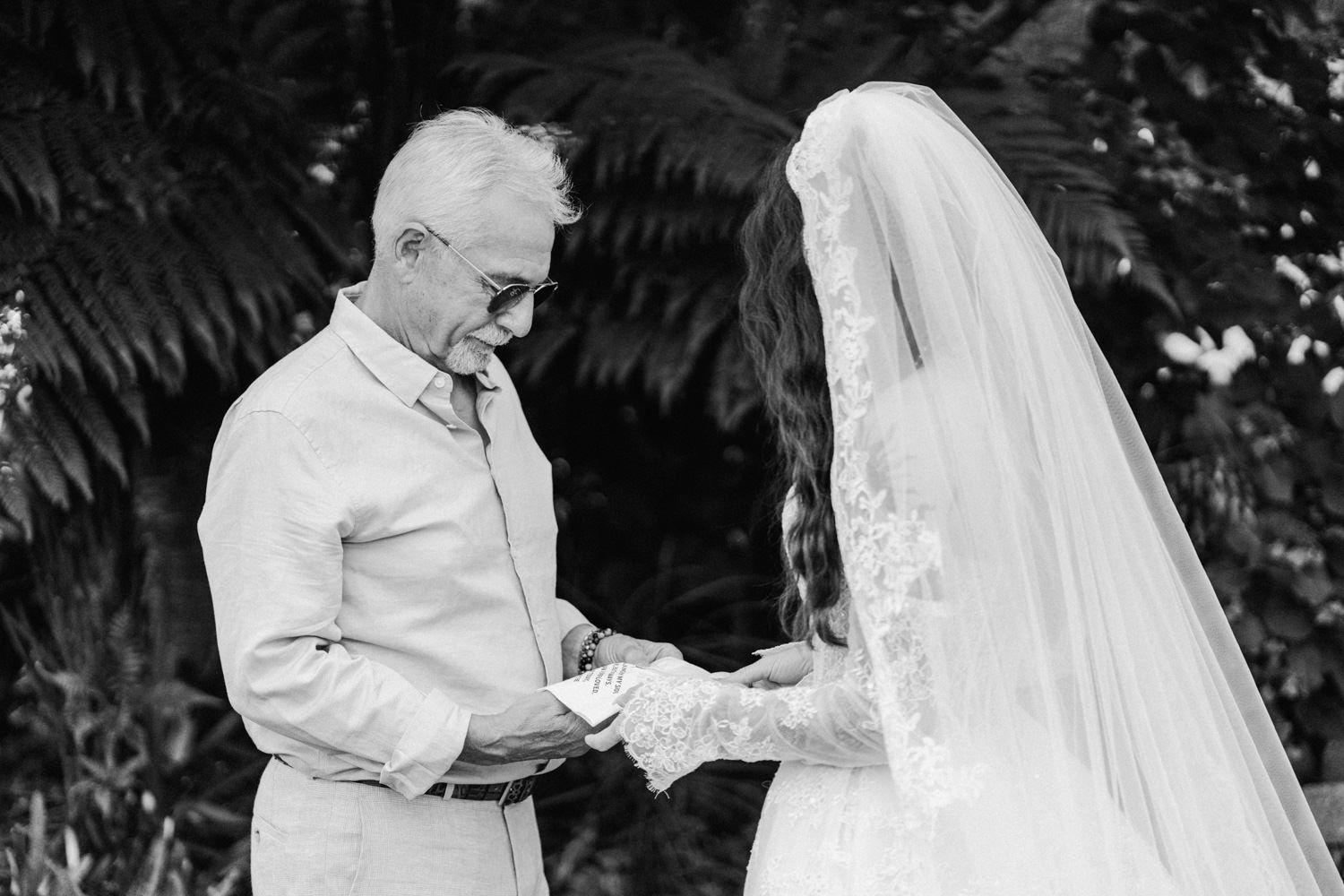 Black and white photo of a bride holding hands with an older man in a garden setting.
