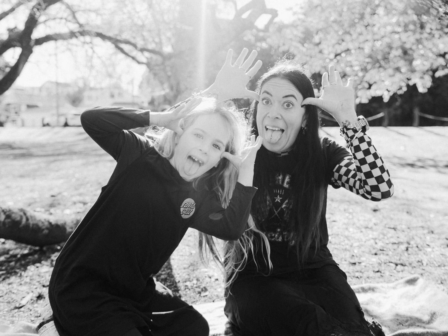 Black and white photo of a girl and woman making silly faces together outdoors in a park.