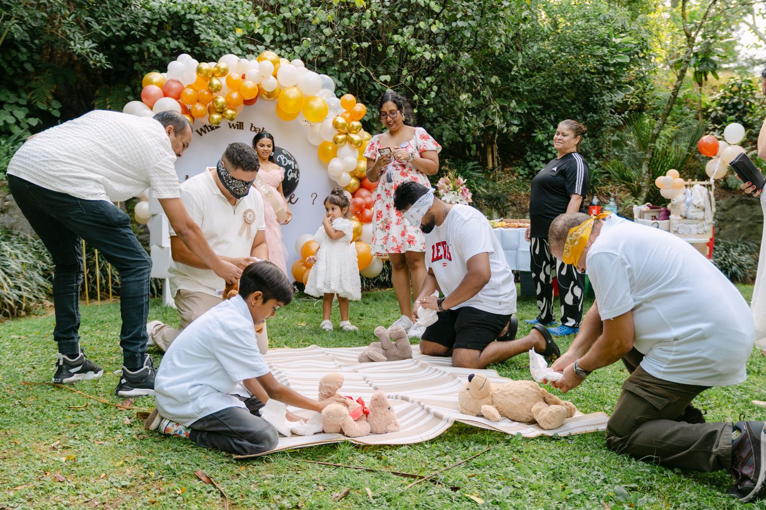 Family and friends gather outdoors at a colorful balloon-decorated birthday party, tending to a baby on a picnic blanket.