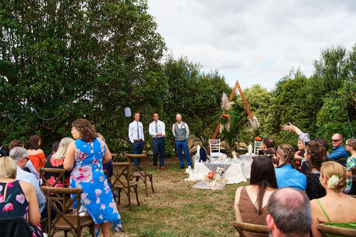 Outdoor wedding ceremony with guests seated on wooden chairs, officiant and groomsmen standing at altar amid lush green trees.