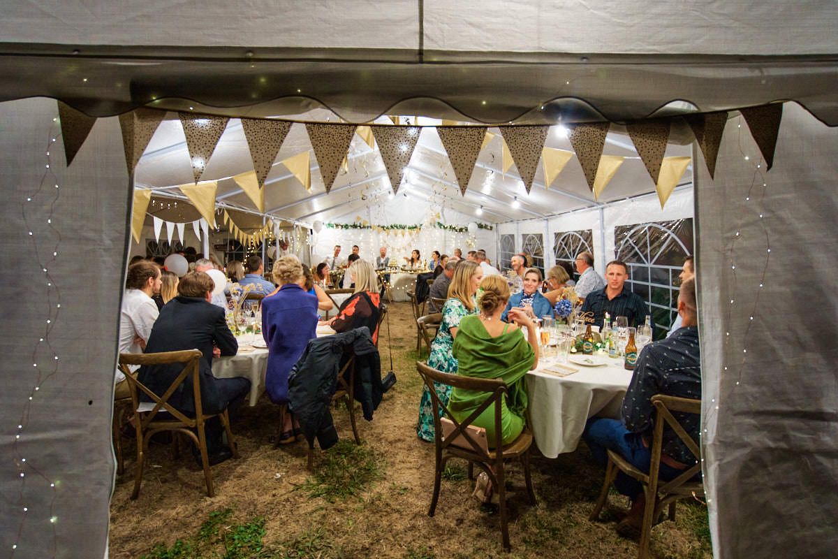 Guests dining under a decorated marquee tent with bunting lights at an outdoor evening wedding reception.