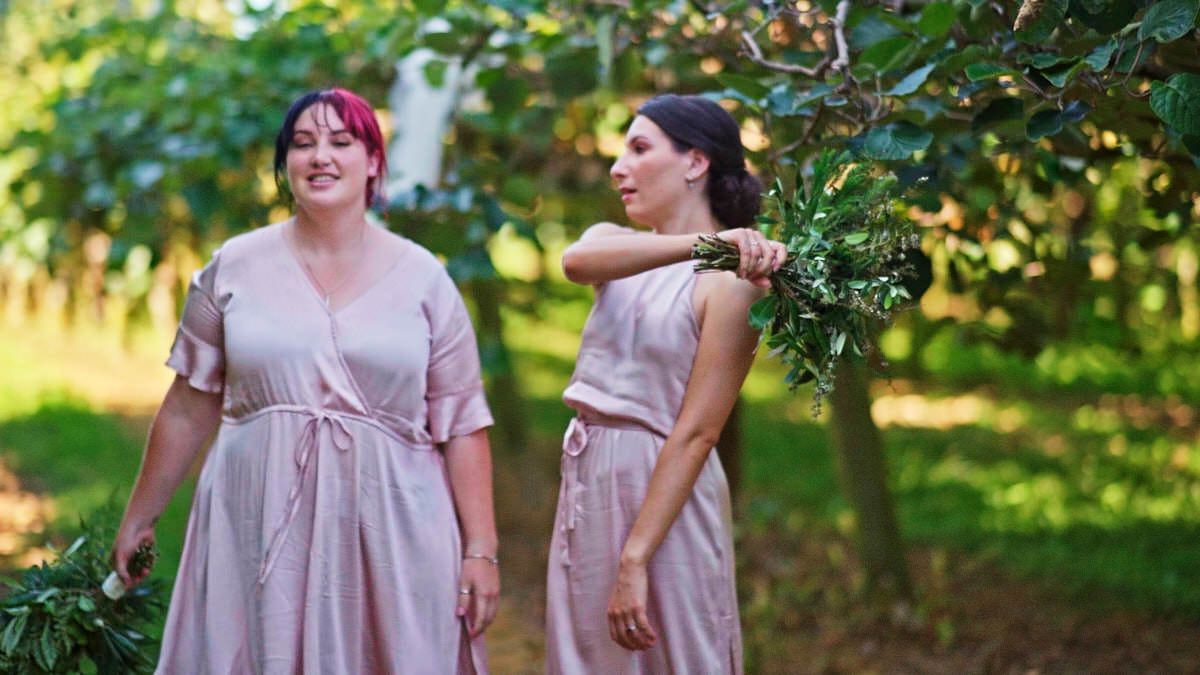 Two bridesmaids in pink dresses walking outdoors carrying greenery bouquets in a lush garden setting.