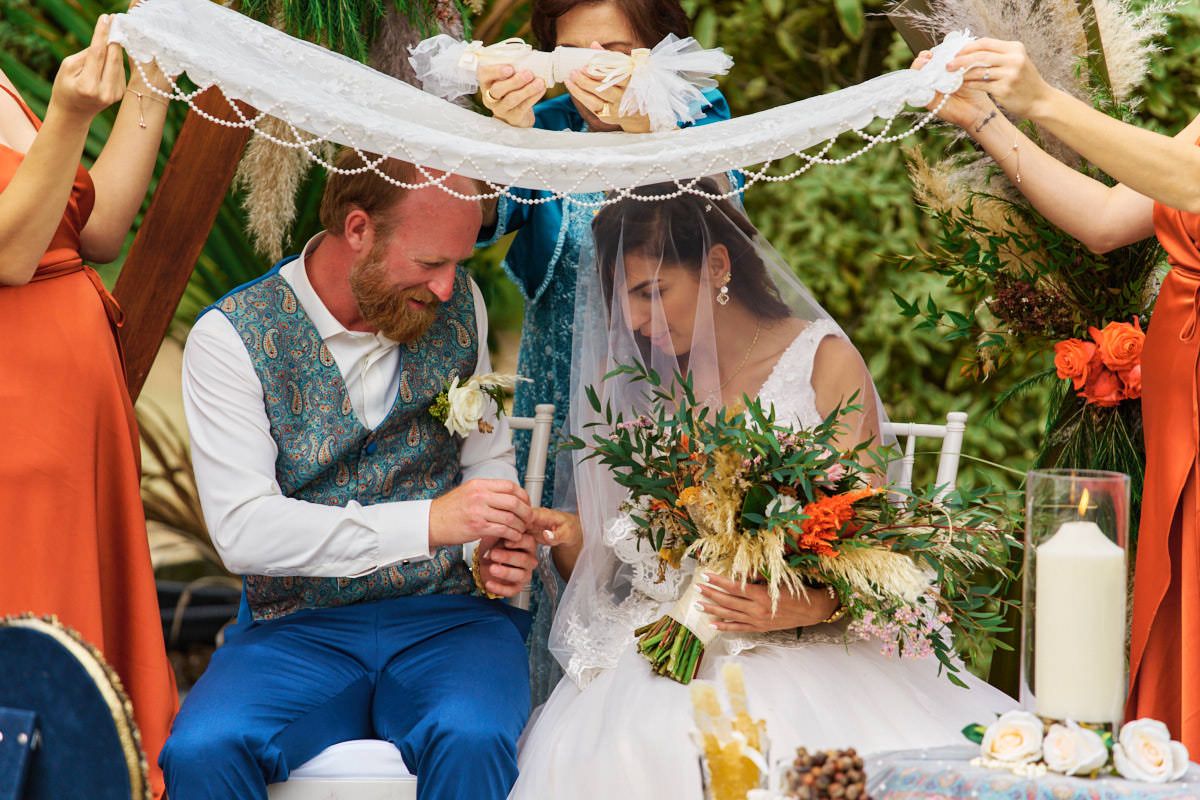 Bride and groom laugh under lace canopy during outdoor wedding ceremony, bride holding rustic bouquet.