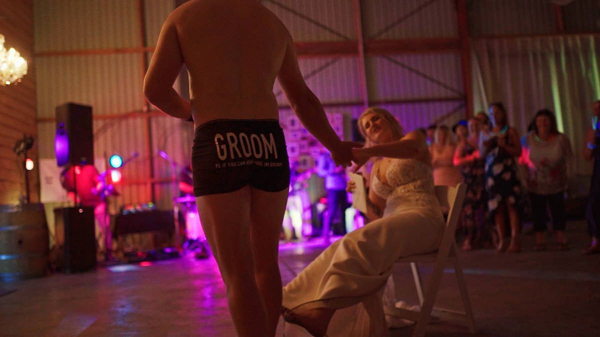 Groom in GROOM shorts dancing for seated bride at lively indoor wedding reception with colorful lighting.