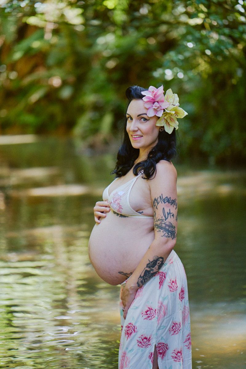 Pregnant woman with floral crown standing in a serene creek, wearing a floral dress, showcasing her baby bump outdoors.