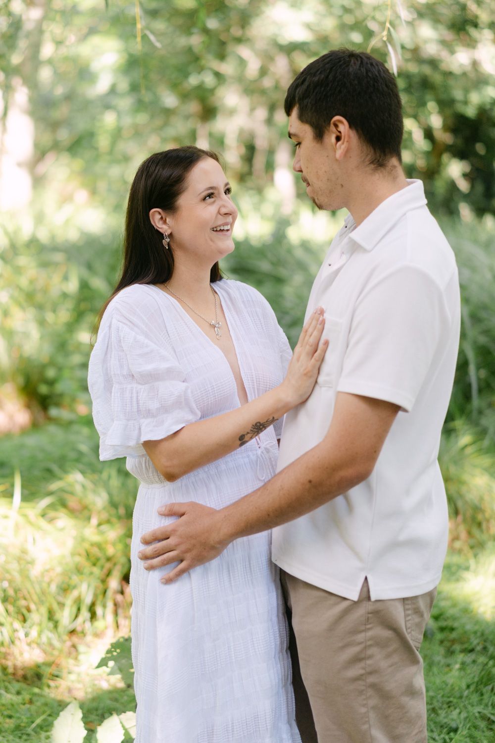 Pregnant couple sharing a tender moment outdoors, woman in white dress smiling at man in lush green garden setting.