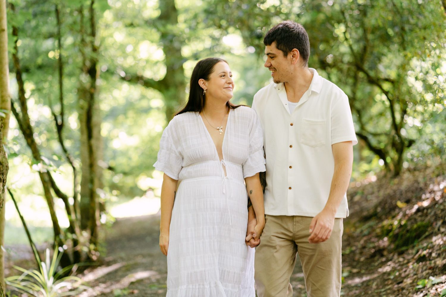 Couple holding hands and smiling at each other while walking on a forest path, woman in white dress.