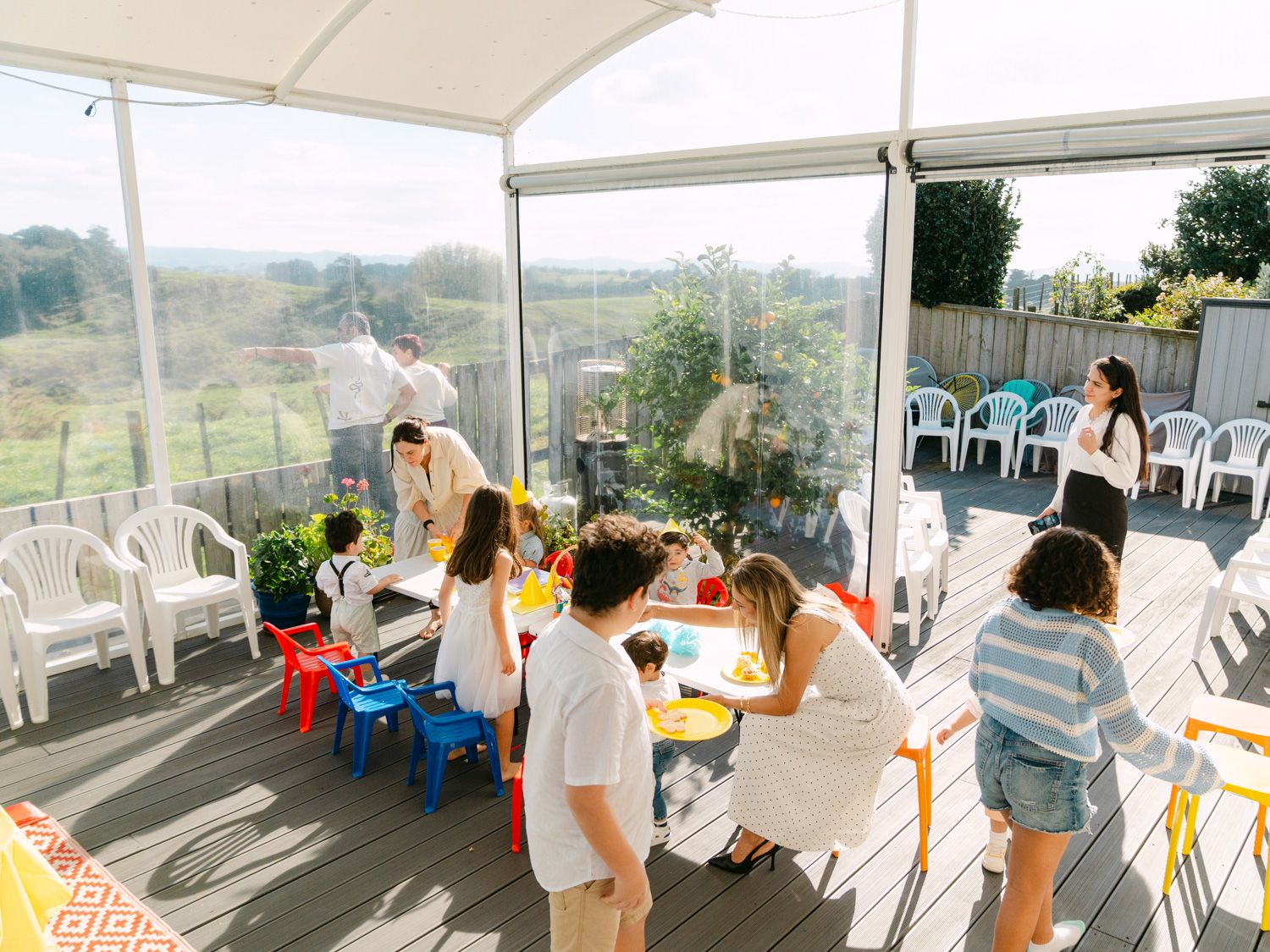 Children and adults gather on a sunny outdoor deck with colorful chairs and panoramic garden views.
