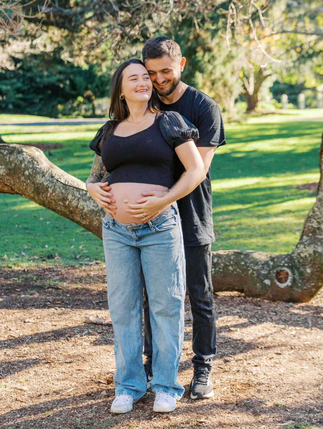 Happy couple in a park, man embracing pregnant woman showing baby bump, both wearing black tops and jeans.