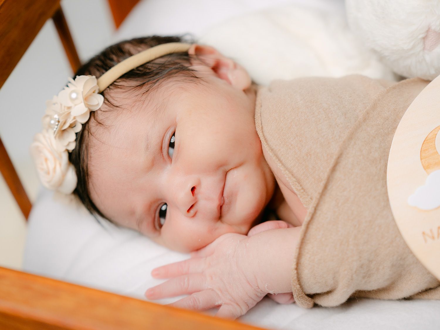 Close-up of a newborn baby wearing a floral headband, wrapped in a beige swaddle, smiling softly.