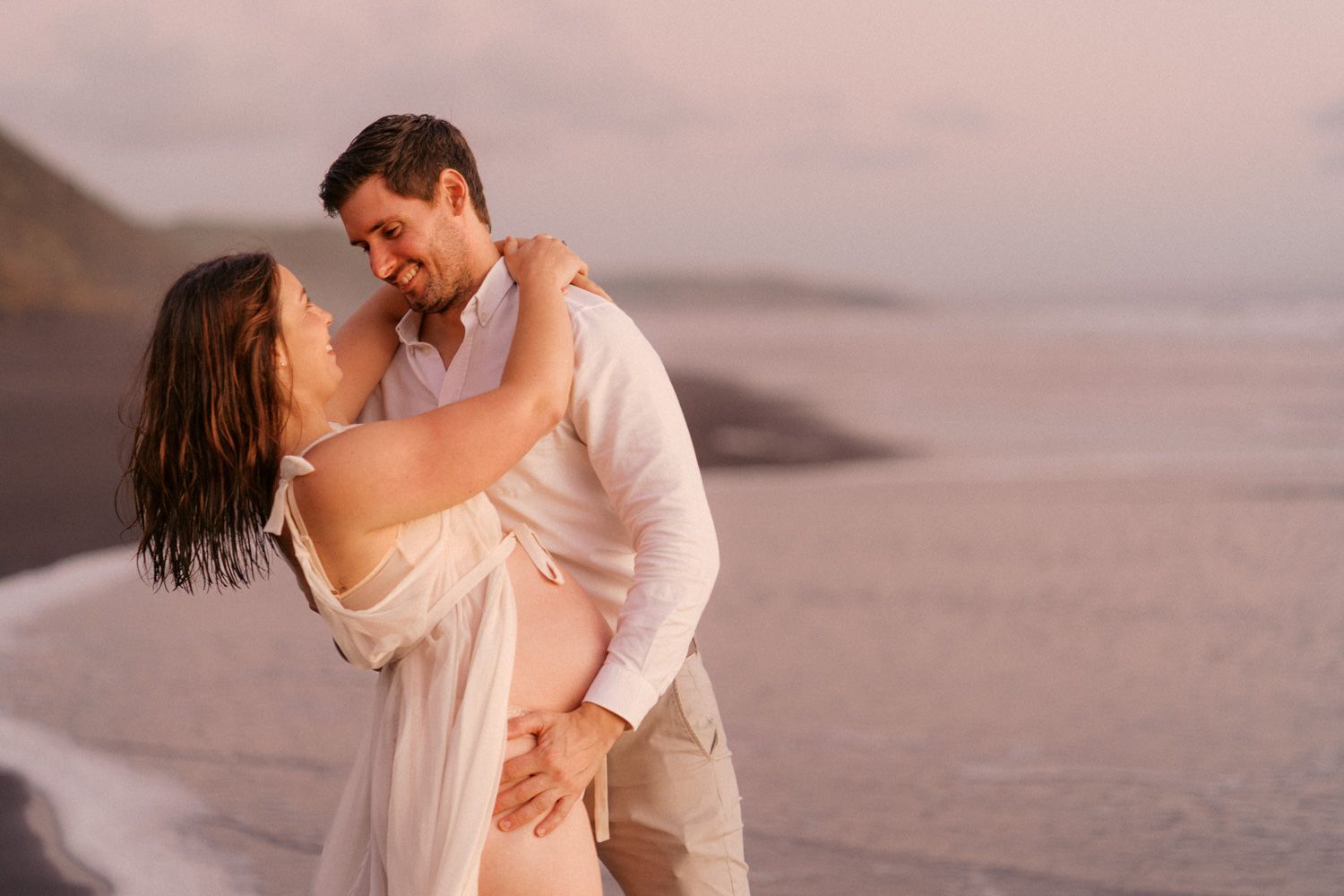 Couple embracing romantically on a beach at sunset, woman in flowing white dress, man in light outfit.