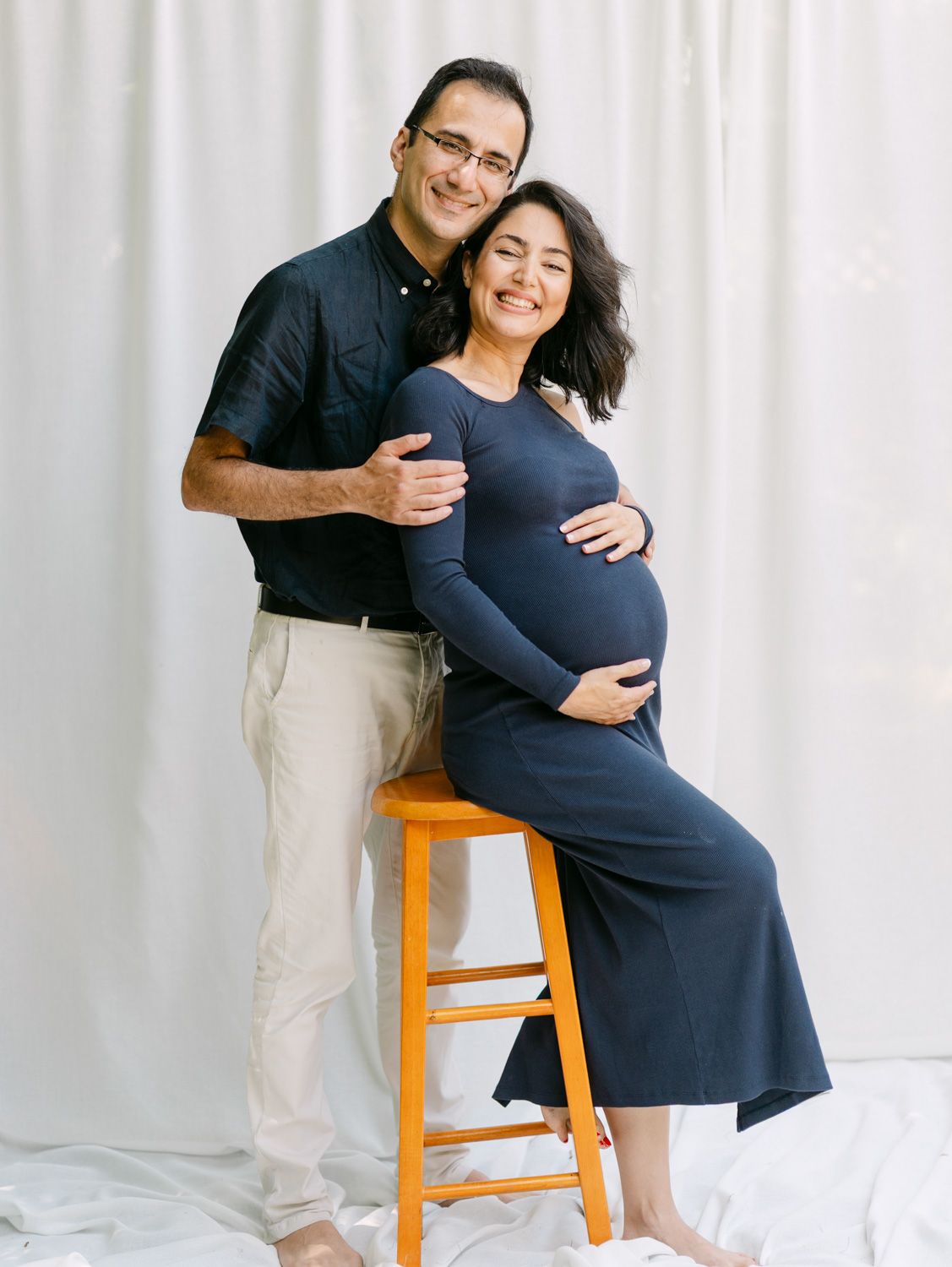Smiling pregnant woman in navy dress sits on stool while partner embraces her in a bright studio maternity photo.