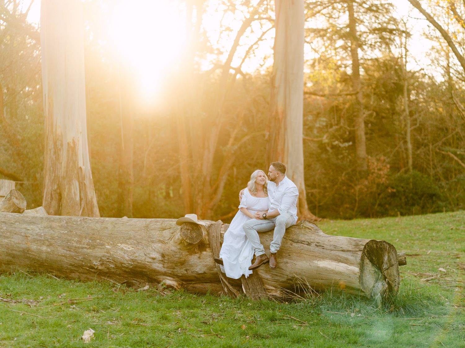 Couple sitting on a fallen log at golden hour sunset in a lush green park, bathed in warm glowing light.
