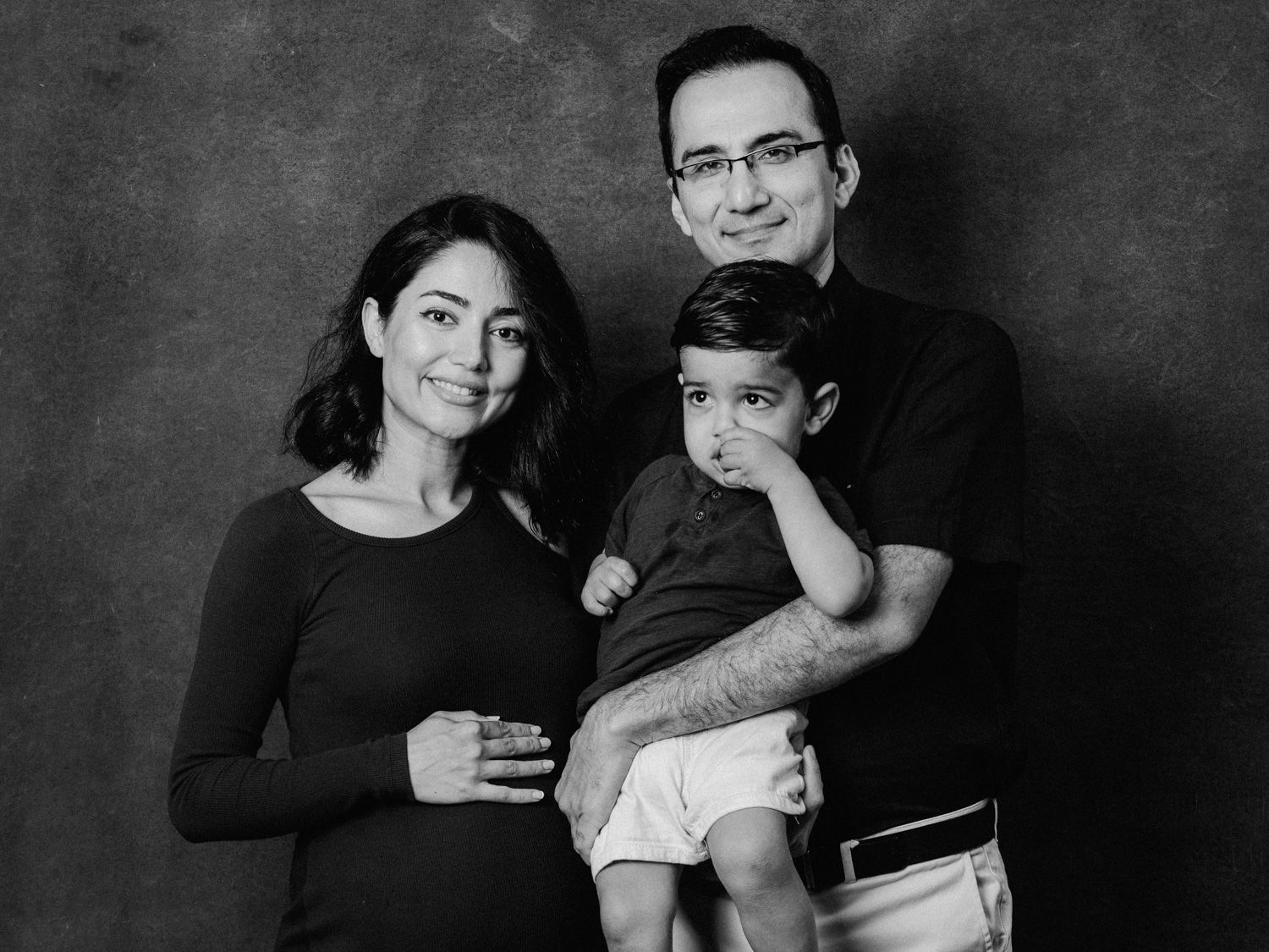 Black and white family portrait of pregnant woman, man with glasses, and young toddler boy posing together.