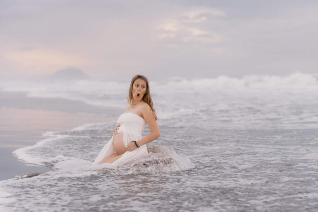 Pregnant woman in white dress sitting in ocean waves at sunset, looking surprised by the water.