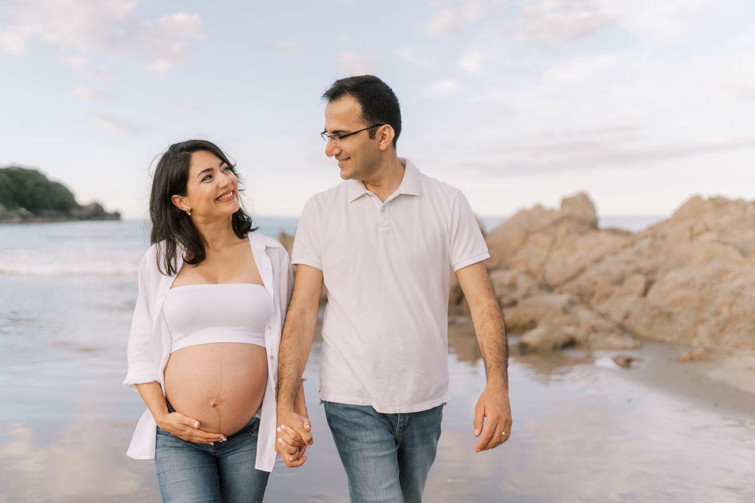 Pregnant couple holding hands smiling at each other on a beach during golden hour maternity photoshoot.