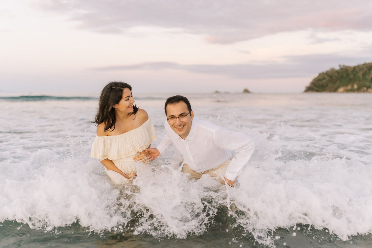 Couple laughing and playing in ocean waves at sunset beach, wearing white outfits during romantic engagement photo session.