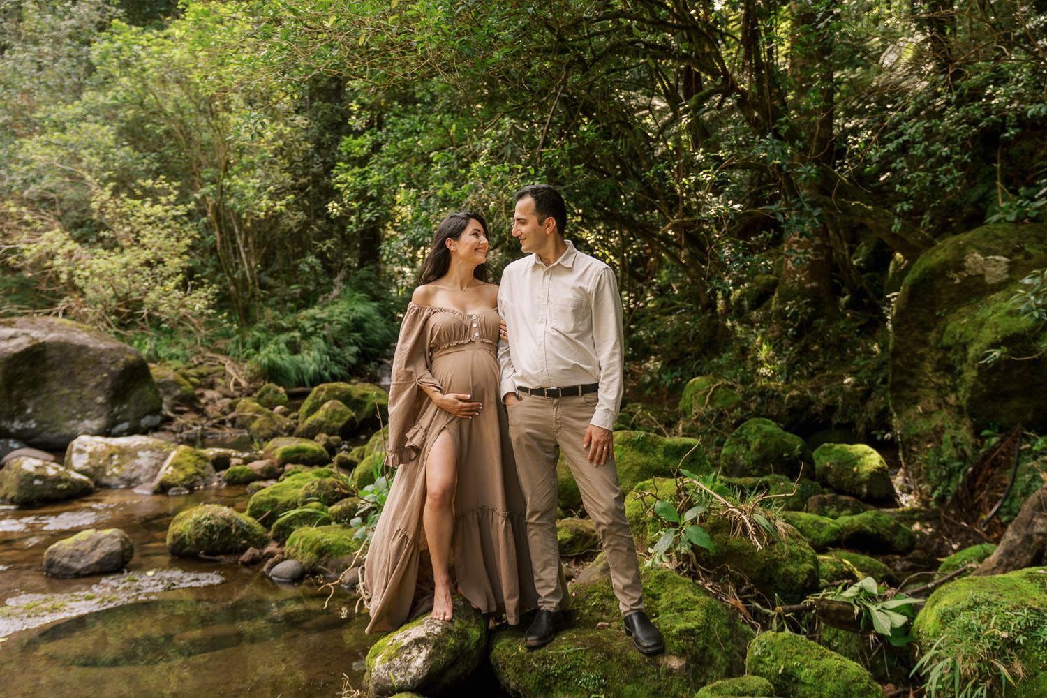 Pregnant couple standing on mossy rocks by a forest stream, surrounded by lush green trees in a serene nature setting.