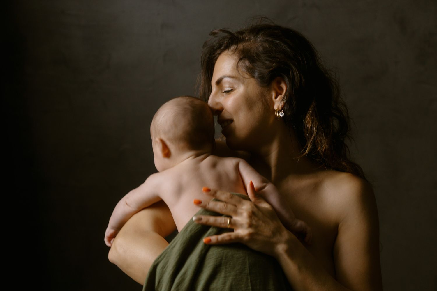 A mother tenderly holds her newborn baby against her bare shoulder in a softly lit, intimate portrait.