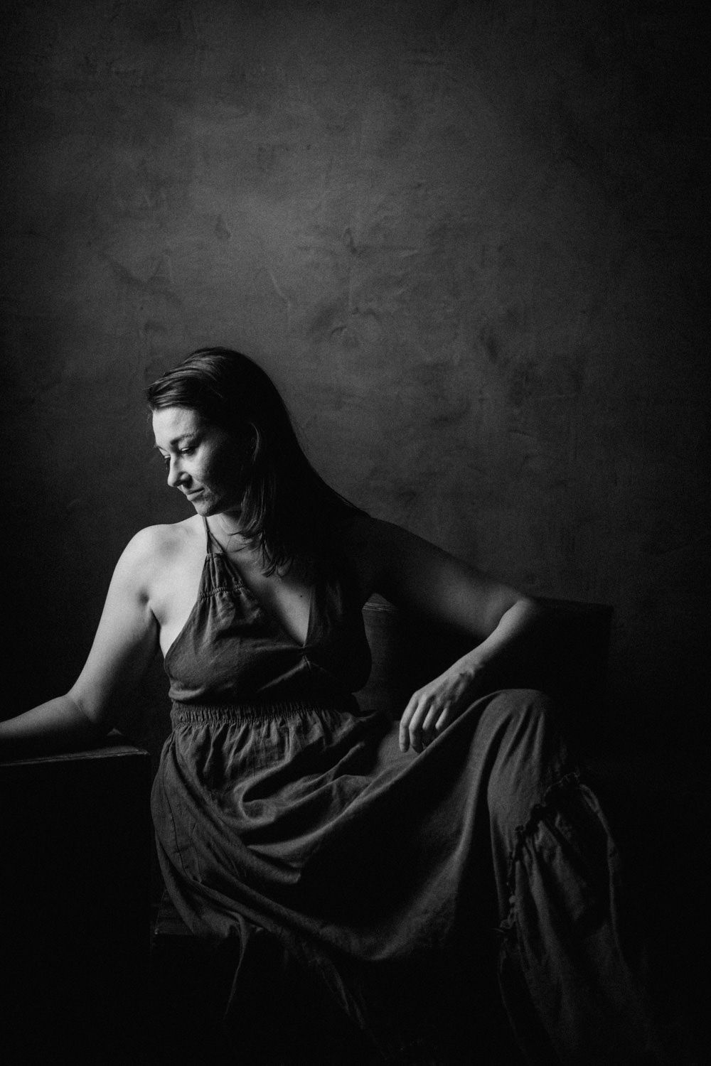 Black and white portrait of a woman in a halter dress seated gracefully against a dark textured studio background.