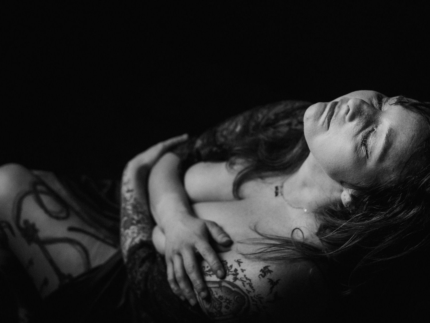 Black and white photo of a tattooed woman reclining with eyes closed in dramatic low-key lighting.