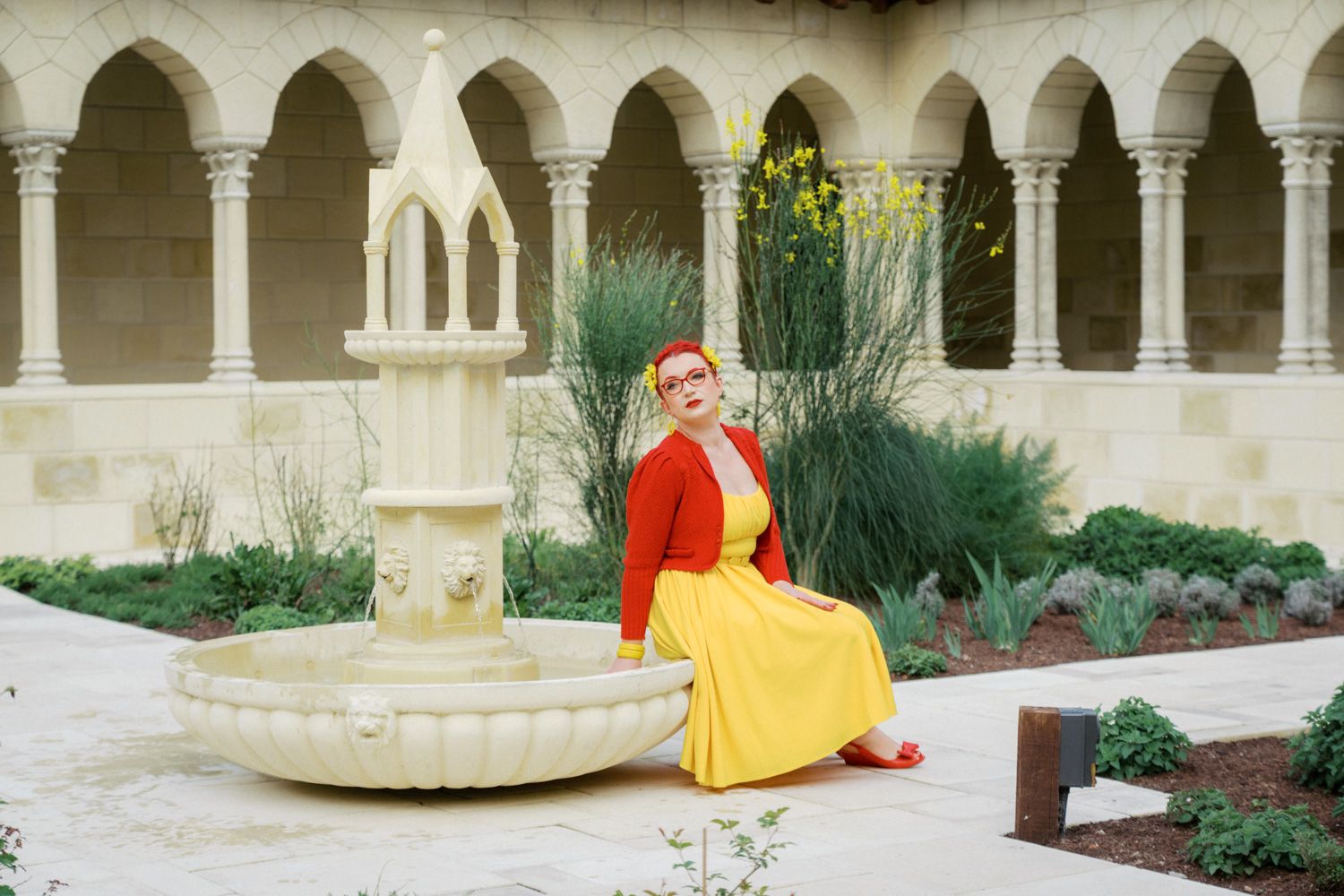 Woman in yellow dress and red cardigan sits by stone fountain in elegant arched courtyard garden.