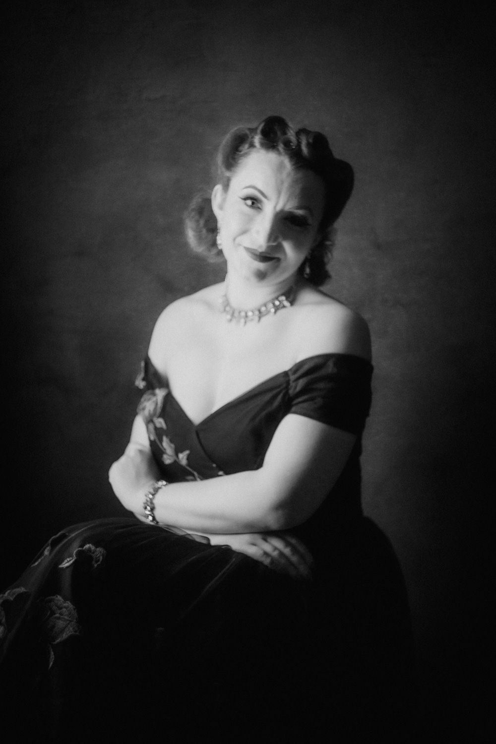 Black and white portrait of an elegant woman in an off-shoulder gown and diamond necklace, posing gracefully.