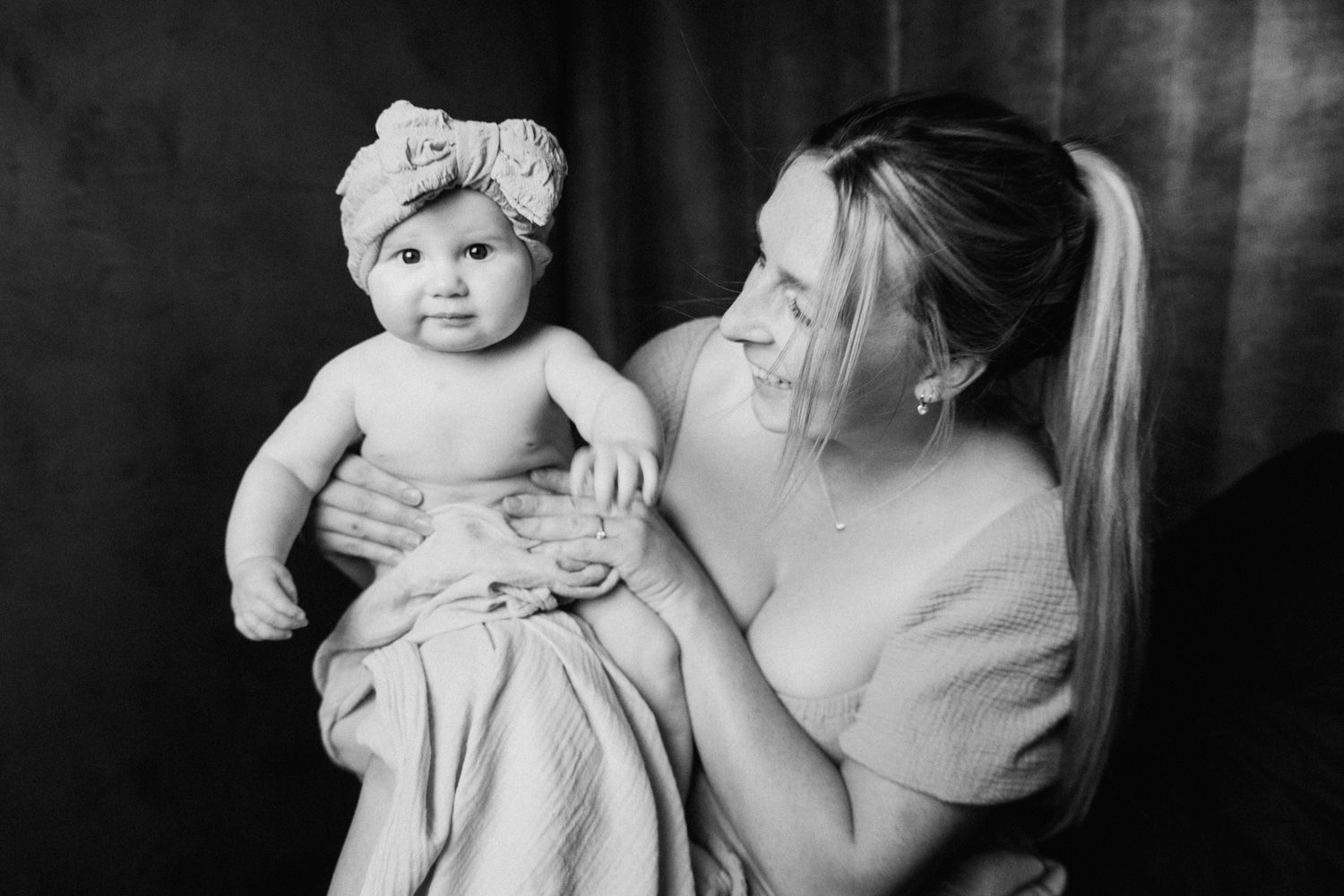 Black and white photo of a smiling mother holding her baby girl wearing a floral headband wrapped in a soft blanket.