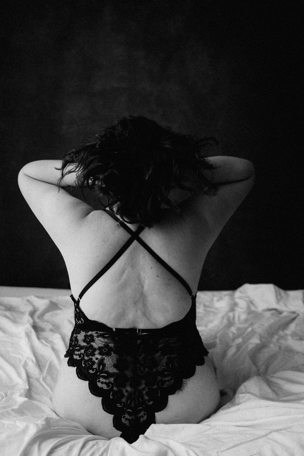 Black and white boudoir photo of a woman in lace lingerie sitting on white bed sheets, back facing camera.