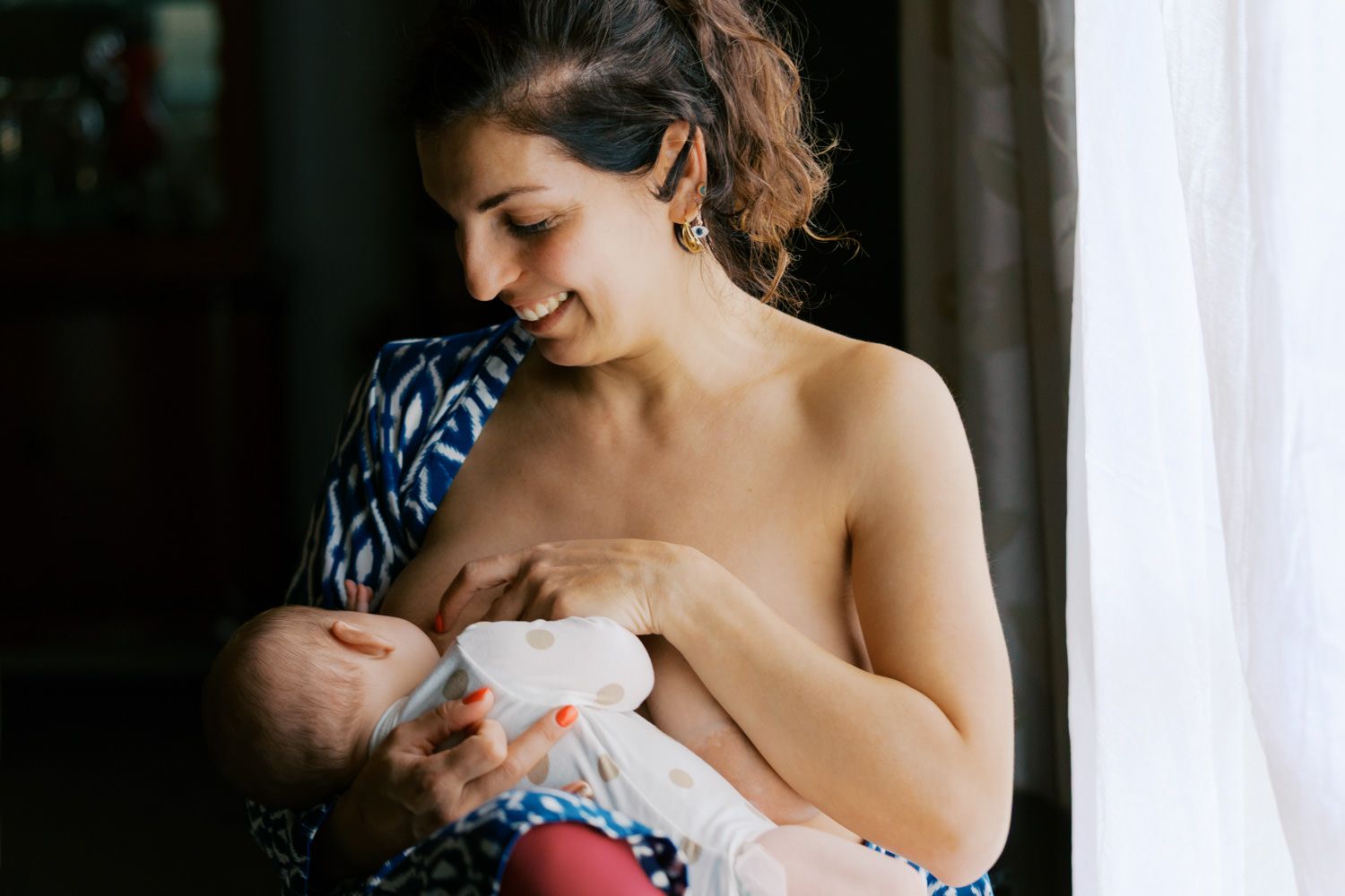 Smiling mother breastfeeding her newborn baby while standing near a window in a blue patterned wrap.