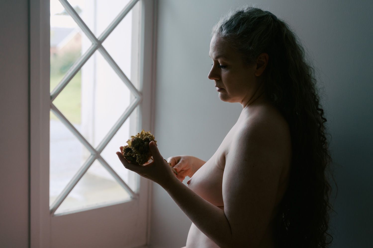 Woman with curly hair standing by a window holding a small object in soft natural light.