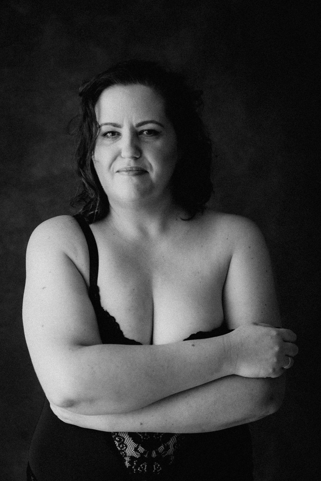Black and white portrait of a confident woman with crossed arms wearing a black lace top against dark background.